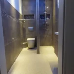 Ein modernes Badezimmer mit grauen Fliesen, einer blauen LED-Decke und Einbauleuchten. Es verfügt über eine wandmontierte Toilette, ein Waschbecken mit Unterschrank und eine ebenerdige Dusche mit Regenduschkopf. An der linken Wand sind Regale mit gefalteten Handtüchern zu sehen.