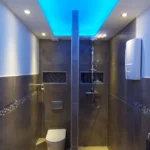 Modernes Badezimmer mit elegantem Design, dunkel gefliesten Wänden und einer Decke mit blauer Umgebungsbeleuchtung. Der Raum verfügt über eine Wandtoilette, einen Duschbereich mit Regenduschkopf, eine kleine Nische für Toilettenartikel und Einbauleuchten in der Decke.