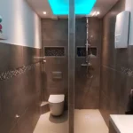 Ein modernes Badezimmer mit futuristischem Ambiente und blau beleuchteter Decke. Der Raum verfügt über eine wandmontierte Toilette, einen durch eine Glaswand abgetrennten Duschbereich und moderne Armaturen. Wände und Boden sind mit glatten, grauen Fliesen bedeckt.