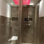 Ein modernes Badezimmer mit braun gefliesten Wänden und beigen Bodenfliesen sowie einer rosafarbenen Decke mit Hintergrundbeleuchtung. Das Badezimmer verfügt auf der einen Seite über eine wandmontierte Toilette und auf der anderen Seite über einen Duschbereich mit Regenduschkopf, abgetrennt durch eine geflieste Trennwand.