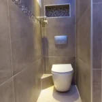 Ein modernes Badezimmer mit grau gefliesten Wänden und Boden. Der Raum verfügt über eine wandmontierte Toilette unter einem eingebauten Regal mit Mosaikfliesendetails. Ein Oberlicht erhellt den Bereich mit natürlichem Licht und unterstreicht das elegante, zeitgenössische Design.